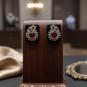 May include: A pair of elaborate earrings on a wooden stand. Each earring has a central red gemstone, surrounded by smaller clear stones and gold-coloured metal. The earrings are displayed against a black background.