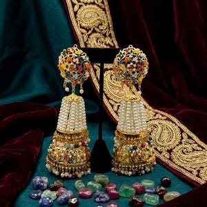 May include: A pair of ornate gold-toned earrings with colorful bead accents and dangling pearl strands. The earrings feature a dome-shaped top and a bell-shaped bottom, displayed against a teal and burgundy backdrop.