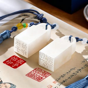 Könnte beinhalten: Zwei weiße, rechteckige Stempel mit geschnitzten chinesischen Schriftzeichen, jeweils an einer blauen Kordel mit Quaste befestigt. Rote Tintenstempel mit chinesischen Schriftzeichen sind ebenfalls sichtbar. Die Stempel liegen auf Papier mit Kalligraphie.