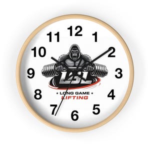 Puede incluir: Un reloj de pared redondo con marco de madera clara. La esfera blanca presenta números negros, un gorila levantando pesas y el texto "LSL LONG GAME LIFTING". Las manecillas del reloj son negras.