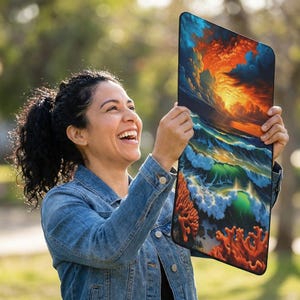 Pode incluir: Uma obra de arte retangular que retrata uma cena oceânica dinâmica com ondas quebrando, um pôr do sol ardente e corais. A obra de arte é segurada por uma pessoa vestindo uma jaqueta jeans.