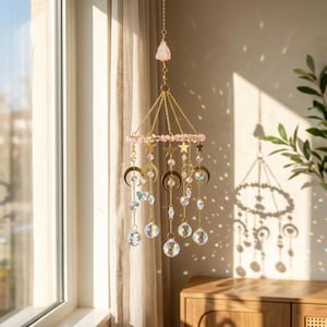 May include: A hanging suncatcher featuring a gold-toned metal frame with dangling chains, clear crystal spheres, and small gold-toned moon and star charms. A row of light pink beads encircles the top, topped with a raw pink crystal point.