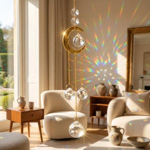 May include: A gold-toned metal crescent moon suncatcher featuring a face design, suspended by a delicate chain. The ornament includes multiple clear, faceted crystal prisms that refract light, creating colorful rainbow patterns on the wall.