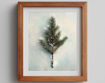 May include: A framed print titled Annaive Studio Prints featuring a single green pine branch with light dusting of white snow against a pale blue textured background. The artwork is enclosed in a medium-toned wooden frame with a white mat border.