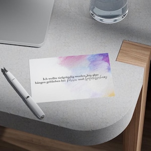 Pode incluir: Uma caneta-tinteiro branca repousa sobre uma mesa cinza clara ao lado de um cartão com arte em aquarela em rosa, roxo e amarelo. O cartão tem texto em alemão. Um copo de água e um laptop também são visíveis.