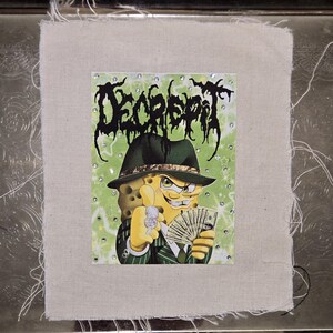 May include: A fabric patch featuring a cartoon character in a green pinstripe suit and fedora, holding cash and a ring. The background is light green with the word "DECREPIT" in black, stylized text. Rhinestones surround the image.