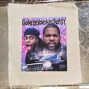 May include: A fabric patch featuring a movie poster with the text "GOREBEYONDNECROPSY". The image shows two men, one wearing a blue cap, and a black car. The background is a pink and purple gradient with sparkling accents.