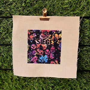 May include: A square artwork with the word "SIEGE" in gold lettering, featuring a colorful skull design. The artwork is attached to a beige fabric with a gold clip.