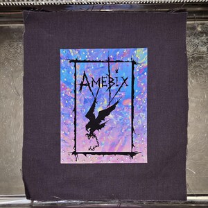 May include: A patch featuring the word "AMEBIX" in a stylized font, with a black bird graphic and chain. The design is set against a colorful, iridescent background and framed with a black border and small, clear embellishments. The patch is on a dark purple fabric.
