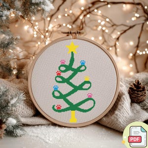 Pode incluir: Um desenho de ponto cruz de árvore de Natal num bastidor de bordado de madeira. A árvore verde é decorada com pegadas em vermelho, azul e rosa, encimada por uma estrela amarela. O fundo é um tecido neutro, com um ícone PDF no canto inferior direito.