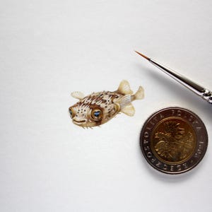 Könnte beinhalten: Eine detaillierte Aquarellmalerei eines kleinen, stacheligen Kugelfisches mit braunen und weißen Markierungen, blauen Augen und einem lächelnden Gesichtsausdruck. Ein feiner Pinsel und eine polnische 2-Złoty-Münze deuten auf den Maßstab des Kunstwerks hin.