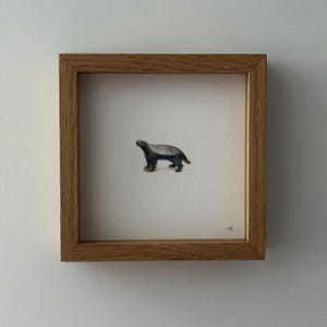 May include: A framed watercolour painting of a badger. The badger is shown in grey and black, with a light brown head and feet. The artwork is set against a white background and framed in a natural wood frame.