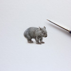 Op de afbeelding: Een gedetailleerd miniatuurschilderij van een grijze wombat in profiel, uitgevoerd met fijne penseelstreken. Een dun, puntig penseel met een donkere steel is diagonaal in de rechterbovenhoek van het beeld geplaatst.