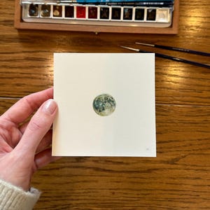 Può includere: Un piccolo dipinto ad acquerello della luna, in tonalità di verde e grigio, centrato su un foglio di carta bianco quadrato. Il dipinto è tenuto in mano, con una tavolozza di acquerelli in legno e pennelli sullo sfondo.