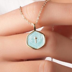 Op de afbeelding: Een goudkleurige ketting met een zeshoekige hanger. De hanger heeft een lichtblauwe emaille achtergrond met een klein kruis in het midden. De ketting wordt door een persoon vastgehouden.