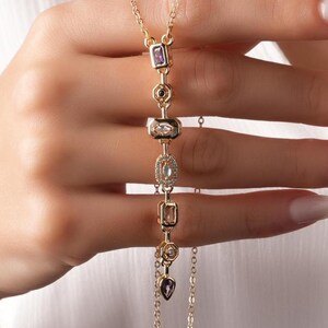 Puede incluir: Un collar dorado con una serie de dijes decorativos. Los dijes incluyen una piedra rectangular púrpura, una piedra circular negra, una piedra rectangular transparente, una piedra ovalada transparente y una piedra en forma de corazón púrpura. El collar es sostenido por una persona.