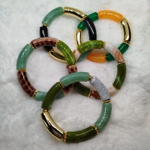Peut inclure: Plusieurs bracelets de perles colorées. Les bracelets présentent des perles vertes, orange, marron et dorées. Les perles sont reliées par des accents métalliques dorés. Les bracelets sont disposés en superposition, créant un effet de superposition.