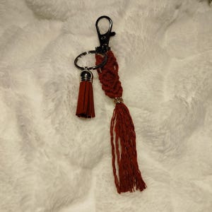 Peut inclure: Un porte-clés en macramé bordeaux avec un pompon et un fermoir en métal. Le porte-clés présente un motif tissé et un petit accent doré. Le pompon est fait d'un matériau doux semblable à du daim.