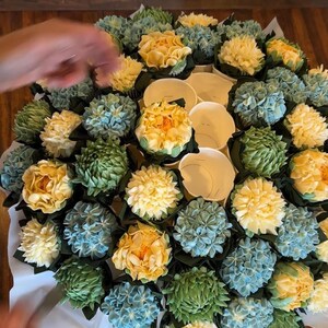 May include: A bouquet of cupcakes decorated to resemble flowers. The cupcakes are arranged in a circular shape, with yellow, blue, and green frosting. The cupcakes are in white paper cups.