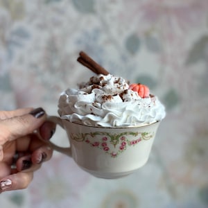 Puede incluir: Una taza de té de porcelana de estilo vintage rellena de nata montada blanca, decorada con una rama de canela marrón y una pequeña calabaza naranja. La taza presenta un delicado estampado floral con rosas rosadas y hojas verdes sobre fondo crema.