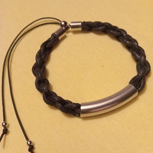 Może przedstawiać: Czarna pleciona bransoletka ze srebrnymi metalowymi detalami. Bransoletka ma pleciony wzór i regulowane czarne sznurki ze srebrnymi koralikami. Prezentowana jest na żółtej powierzchni.