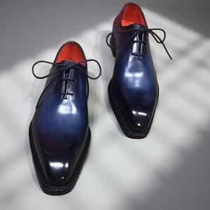 May include: A pair of men's dress shoes with a gradient blue and black finish. The shoes have a pointed toe, a lace-up closure, and a red interior. The shoes are made of a glossy material.
