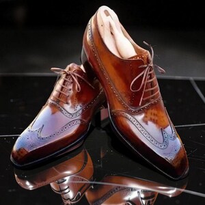 Può includere: Un paio di scarpe eleganti in pelle marrone con dettagli brogue e lacci marroni chiari. Le scarpe hanno una finitura lucida e si riflettono sulla superficie scura. Il design classico è visibile.