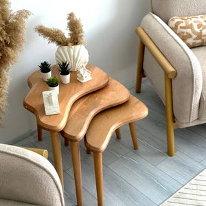 May include: A set of three nesting tables with organic, kidney-shaped wooden tops and tapered legs. The tables are arranged in a staggered formation, featuring a light brown wood grain finish. Each table stands on three cylindrical wooden legs.