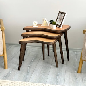 May include: A set of three nesting tables with kidney-shaped tops featuring light wood surfaces and dark brown edges. Each table has three tapered dark brown wooden legs. The tables are stacked together, with small decorative items on top.