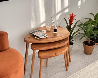 May include: A set of three nesting tables crafted from light brown wood with smooth, rounded, organic-shaped tops and tapered legs. The tables are stacked in a descending arrangement, with books and a glass fragrance diffuser placed on the largest top.
