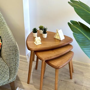 May include: A set of three nested wooden tables with triangular tops and rounded edges. The tables feature a light brown wood grain finish and tapered legs. Three small potted succulents and two white house-shaped ornaments sit on the top table.