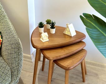 May include: A set of three nested wooden tables with triangular tops and rounded edges. The tables feature a light brown wood grain finish and tapered legs. Three small potted succulents and two white house-shaped ornaments sit on the top table.