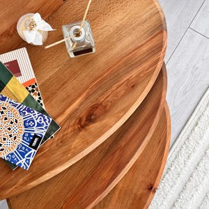 May include: A set of three wooden, oval-shaped nesting tables with a warm, reddish-brown finish. A small glass diffuser with reeds and a decorative candle sit on the top table. A stack of colourful magazines adds a decorative touch.
