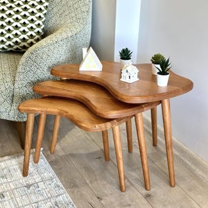 May include: Three wooden nesting tables in a warm, natural wood tone, featuring a distinctive wavy design. Each table has slender, tapered legs. The top table is decorated with small potted plants and miniature houses. The tables are a stylish addition to any living space, and are approximately 76 cm tall.