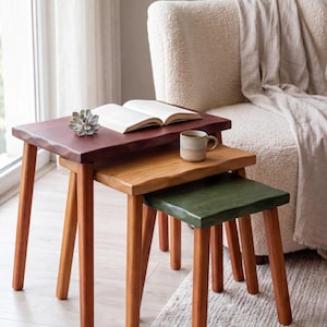 May include: A set of three nesting tables with natural wood legs and rectangular tops in dark brown, medium brown, and dark green. The tables feature live edges and are arranged in a cascading formation, with an open book and a mug on top.