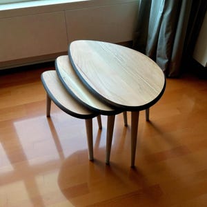 May include: A set of three nesting tables with light wood oval tops and black trim. The tables have light-colored, tapered legs. The tables are arranged in a stacked configuration, with the largest table on top.