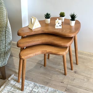 May include: A set of three nested tables made of light brown wood with organic, kidney-shaped tops and tapered legs. The tables are arranged in a tiered stack, with three small potted green succulents and two miniature house figurines on top.