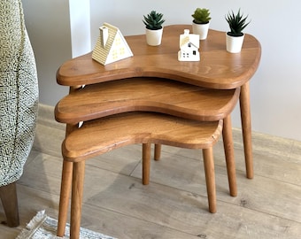 May include: A set of three nested tables made of light brown wood with organic, kidney-shaped tops and tapered legs. The tables are arranged in a tiered stack, with three small potted green succulents and two miniature house figurines on top.
