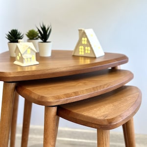 Könnte beinhalten: Ein Satz von drei Holz-Nesting-Tischen mit dreieckiger Oberseite und sich verjüngenden Beinen. Die Tische sind warmbraun und in einer gestapelten Konfiguration angeordnet. Miniatur-Hauslichter und kleine Topfpflanzen befinden sich auf dem oberen Tisch.