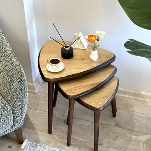 Può includere: Un set di tre tavolini a incastro con piani triangolari in legno con finitura a venature di rovere chiaro e bordi marrone scuro. Ogni tavolo è sostenuto da tre gambe affusolate in legno marrone scuro. I tavoli sono impilati per dimensione.