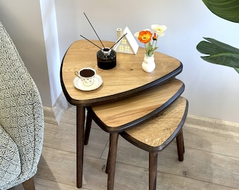May include: A set of three nesting tables with triangular wooden tops featuring a light oak grain finish and dark brown edges. Each table is supported by three tapered dark brown wooden legs. The tables are stacked in a descending size order.