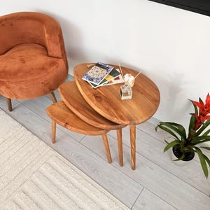 May include: A set of three wooden nesting tables with a natural finish. The tables have an organic shape and are supported by slender, tapered legs. A small orange armchair and a white rug are in the background.