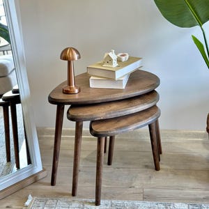 Può includere: Un set di tre tavolini a incastro in legno marrone scuro con una forma triangolare arrotondata e liscia. Ogni tavolo presenta tre gambe affusolate. Una piccola lampada color rame e due libri impilati con un ornamento a casa bianca.