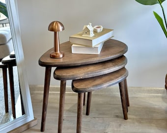 May include: A set of three nesting tables crafted from dark brown wood with a smooth, rounded triangular shape. Each table features three tapered legs. A small copper-coloured lamp and two stacked books with a white house ornament sit on top.