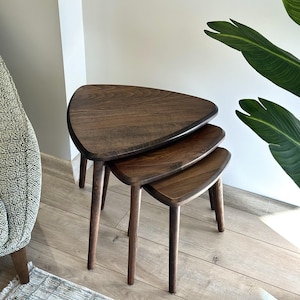 May include: Set of three nesting tables with a triangular top and dark brown finish. The tables have rounded corners and slender, tapered legs. The largest table is approximately 24 inches wide, 20 inches deep, and 18 inches tall.