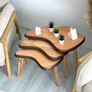 Könnte beinhalten: Ein Set aus drei Satztischen mit organisch geformten, nierenförmigen Holzplatten, die hellbraune Oberflächen und dunkelbraune Kanten aufweisen. Jeder Tisch wird von drei konisch zulaufenden Holzbeinen getragen.