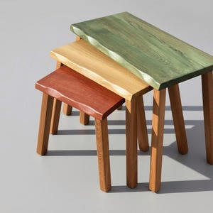 Könnte beinhalten: Dreiteiliges Set von Beistelltischen mit Holzbeinen und rechteckigen Tischplatten. Die Tischplatten sind in Rot-, Gelb- und Grüntönen gebeizt. Die Tische sind so konzipiert, dass sie ineinander passen.