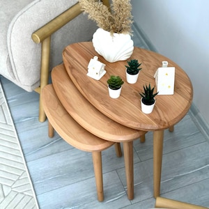 Può includere: Un set di tre tavolini a incastro in legno chiaro con piani ovali lisci e gambe affusolate. I tavolini sono disposti a cascata e ospitano un vaso bianco a forma di conchiglia con erba secca, tre piccole piante grasse in vaso e due ornamenti bianchi a forma di casetta.