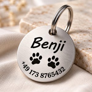 Könnte beinhalten: Eine kreisförmige Haustiermarke aus gebürstetem Metall mit der Gravur Benji, zwei schwarzen Pfotenabdrücken und der Telefonnummer +49 173 8765432, versehen mit einem silbernen Metallring zur Befestigung am Halsband.