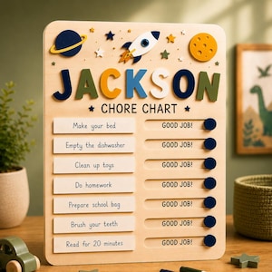 May include: A light-colored wooden chore chart featuring space-themed decorations including a rocket, planet, and moon. The name JACKSON is displayed in large, colorful letters at the top. It includes seven tasks with sliding dark blue buttons.
